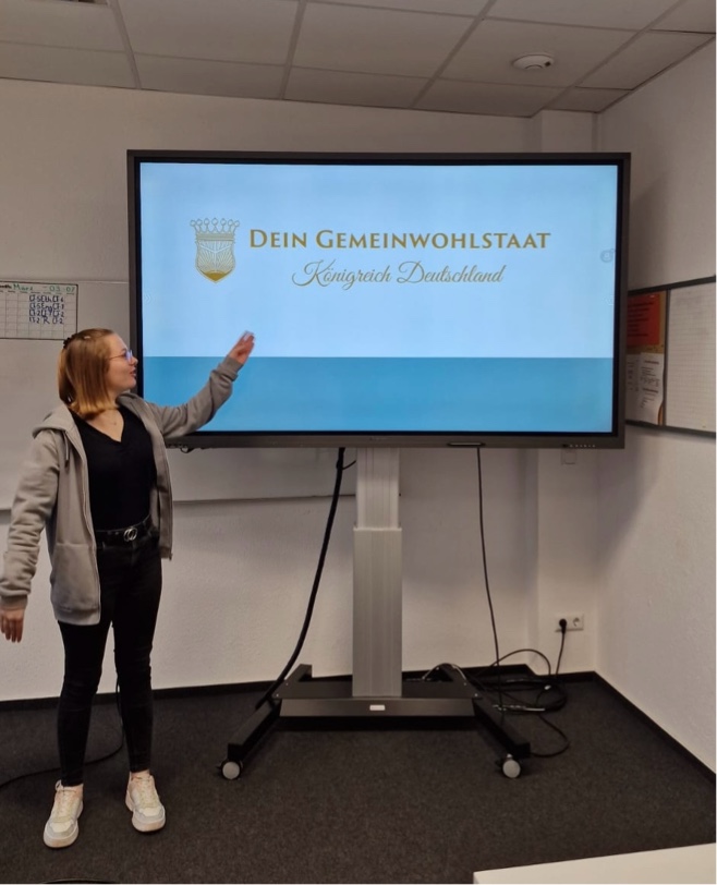 Projektwoche „Umgang mit Verschwörungstheorien" Eine Schülerin steht vor einem großen Bildschirm, auf dem eine Präsentationsfolie mit dem Titel „Dein Gemeinwohlstaat – Königreich Deutschland“ zu sehen ist. Sie zeigt mit der Hand auf den Bildschirm.
