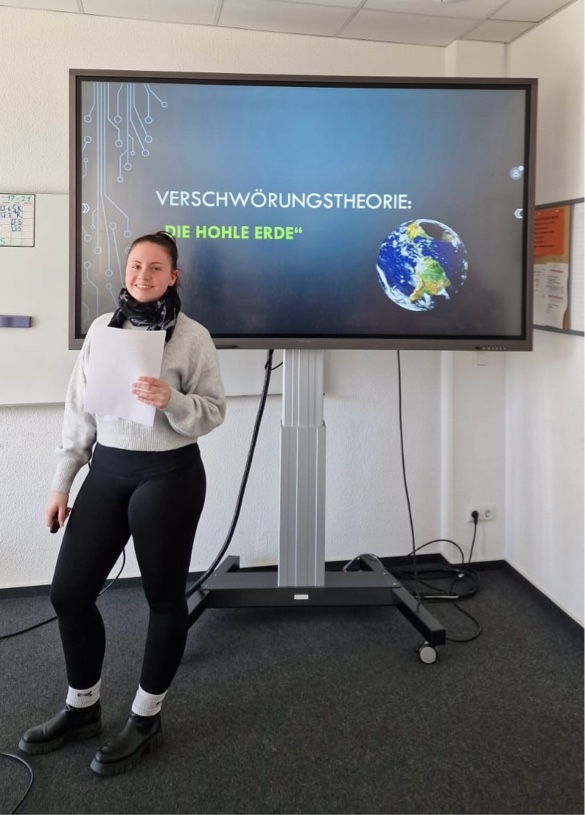 Projektwoche „Umgang mit Verschwörungstheorien" Eine Schülerin steht vor einer Folie zur „Hohle Erde“-Verschwörungstheorie. Auf dem Bildschirm ist ein Bild der Erde mit einem Hohlraum im Inneren zu sehen.