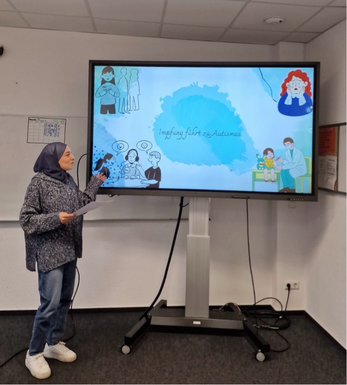Projektwoche „Umgang mit Verschwörungstheorien" Eine Schülerin präsentiert eine Folie mit dem Titel „Impfung führt zu Autismus“, versehen mit Illustrationen von Ärzt:innen, Kindern und Eltern – vermutlich zur Aufklärung über diese Verschwörungstheorie.