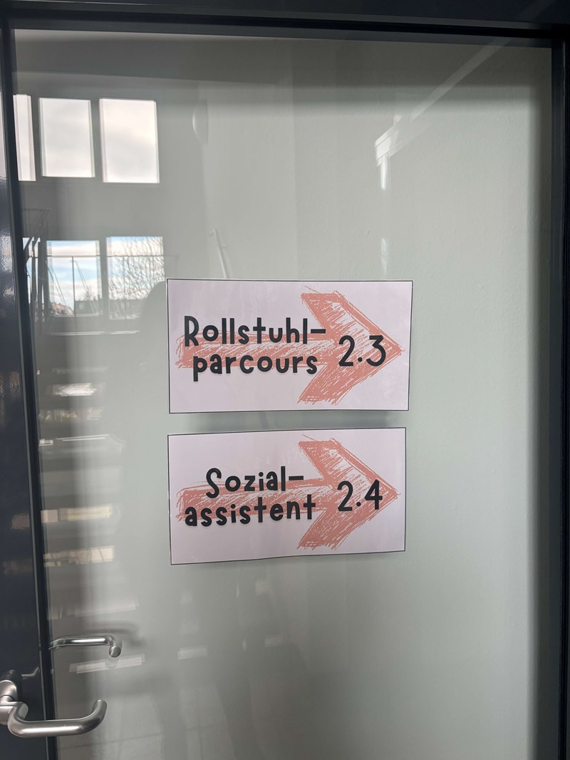 An einer Glastür sind zwei weiße Schilder mit handgeschriebener schwarzer Schrift und jeweils einem großen, rot schraffierten Pfeil nach rechts angebracht. Das obere Schild trägt die Aufschrift 'Rollstuhl-Parcours 2.3', das untere 'Sozialassistent 2.4'. Die Tür ist mit einem silbernen Türgriff ausgestattet, und durch die Glasfläche sind Fenster und ein Teil des Innenraums mit Tageslicht sichtbar. Die Schilder weisen auf Räume oder Stationen im Gebäude der Euro Akademie Zwickau hin, die im Rahmen des Tages der offenen Tür besucht werden können.