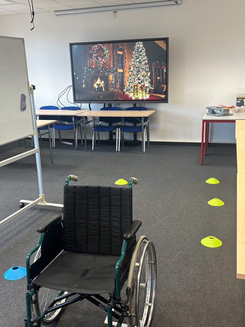 In einem hellen, modernen Seminarraum der Euro Akademie Zwickau steht ein schwarzer Rollstuhl mit grünem Rahmen auf grauem Teppichboden. Im Hintergrund sind mehrere blaue Stühle an einem langen Tisch aufgereiht. An der Wand hängt ein großer Flachbildfernseher, auf dem ein festlich geschmückter Weihnachtsbaum und ein Kaminfeuer zu sehen sind. Auf dem Boden sind mehrere bunte Markierungshütchen in Gelb und Blau verteilt, die offenbar für eine Übung oder einen Parcours genutzt werden. Links im Bild ist ein weißes Flipchart zu erkennen, rechts ein kleiner Tisch mit einigen Gegenständen darauf. Die Szene entstand im Rahmen des Tages der offenen Tür an der Euro Akademie Zwickau.