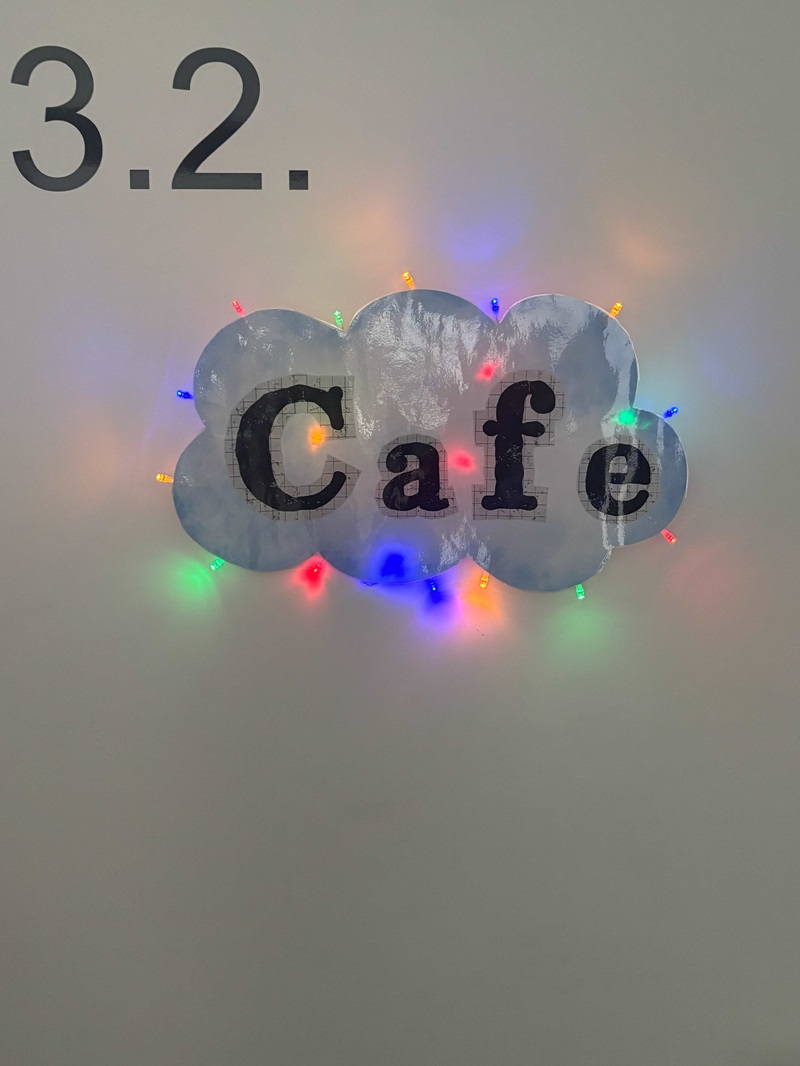 An einer hellen Wand ist die Zahl '3.2.' in schwarzer Schrift zu sehen. Darunter befindet sich ein dekoratives Schild in Form einer Wolke mit dem Wort 'Cafe' in großen, schwarzen Buchstaben. Das Schild ist mit bunten Lichterketten umrandet, die in verschiedenen Farben wie Rot, Grün, Blau, Gelb und Orange leuchten und eine freundliche Atmosphäre schaffen. Das Bild entstand im Rahmen des Tages der offenen Tür an der Euro Akademie Zwickau und zeigt einen Bereich, der als Café gekennzeichnet ist.