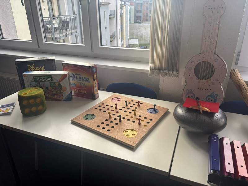 Auf einem weißen Tisch vor einem Fenster mit Blick auf benachbarte Gebäude steht ein Holzspielbrett mit mehreren Löchern und kleinen Stiften, die in einigen Löchern stecken. Links daneben befinden sich drei Spieleschachteln, darunter eine mit der Aufschrift 'Phase' und eine weitere mit dem Titel 'Schule'. Vor den Schachteln liegt ein grüner Würfel mit gelben Punkten. Rechts auf dem Tisch steht ein rundes, schwarzes Musikinstrument, auf dem ein rotes Notenblatt mit einem Holzstab liegt. Daneben liegen drei bunte Metallxylophone in den Farben Blau, Lila und Rot. Im Hintergrund sind weiße Fensterrahmen und vertikale Jalousien zu sehen. Das Bild entstand im Rahmen des Tages der offenen Tür an der Euro Akademie Zwickau.