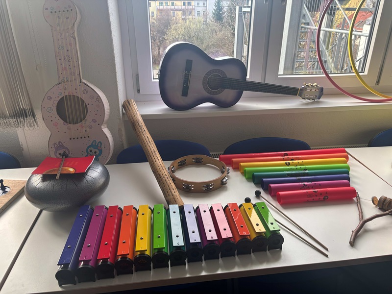 Auf einem weißen Tisch stehen verschiedene Musikinstrumente, darunter ein farbenfrohes Glockenspiel mit Metallplatten in Regenbogenfarben, ein Tamburin mit Holzrahmen und Schellen, sowie ein Holzstab. Daneben liegen zwei Metallklöppel. Im Hintergrund auf der Fensterbank steht eine schwarze Akustikgitarre. Links neben dem Tisch ist eine große, aus Pappe gefertigte Gitarre mit aufgemalten Details zu sehen. Durch das Fenster sind unscharf Bäume und Gebäude zu erkennen. Die Szene wirkt hell und freundlich, typisch für eine Lernumgebung während eines Tages der offenen Tür an der Euro Akademie Zwickau.