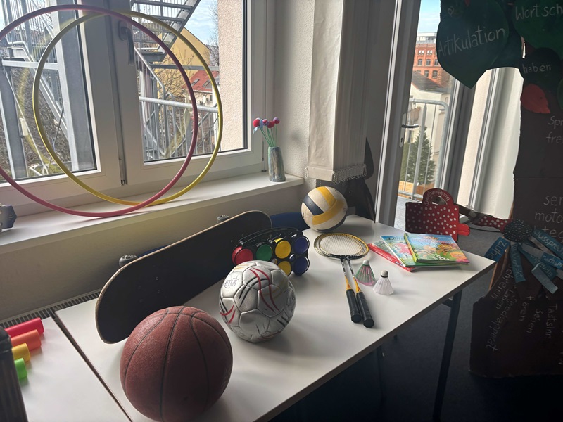 Auf einem weißen Tisch vor einem Fenster mit weißen Rahmen stehen verschiedene Sport- und Spielgeräte. Von links nach rechts sind bunte Hula-Hoop-Reifen an der Wand befestigt, ein Skateboard mit schwarzer Oberfläche, ein brauner Basketball, ein weißer Fußball mit roten Linien, eine Ampelattrappe mit roten, gelben und grünen Lichtern, zwei Badmintonschläger mit weißen Griffen, ein weißer Federball, mehrere bunte Kinderbücher und ein roter Rucksack mit weißen Punkten. Im Hintergrund sind ein Fenster mit Blick auf ein Gebäude und eine weiße Wand zu sehen. Das Bild entstand im Rahmen des Tages der offenen Tür an der Euro Akademie Zwickau.