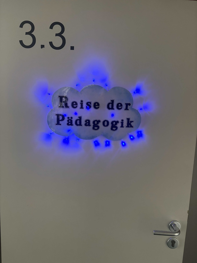 Weiße Tür mit der schwarzen Aufschrift '3.3.' oben links. In der Mitte der Tür befindet sich eine Wolkenform mit der schwarzen Schrift 'Reise der Pädagogik'. Um die Wolke herum leuchten mehrere kleine blaue LED-Lichter, die einen leuchtenden Rahmen bilden. Rechts unten ist ein silberner Türgriff sichtbar. Das Bild entstand im Rahmen des Tages der offenen Tür an der Euro Akademie Zwickau.