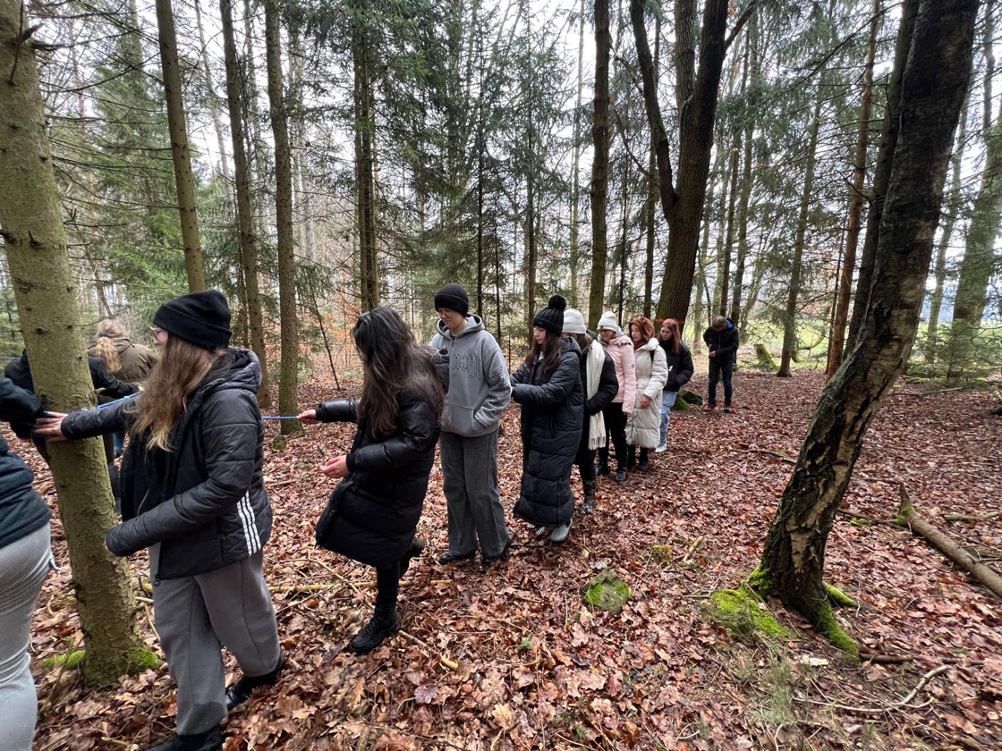 Ein große Gruppe Schüler*innen bewegt sich entlang eines von Baum zu Baum gespannten Seils vorwärts. Die Personen tragen dicke Jacken und teilweise Mützen.