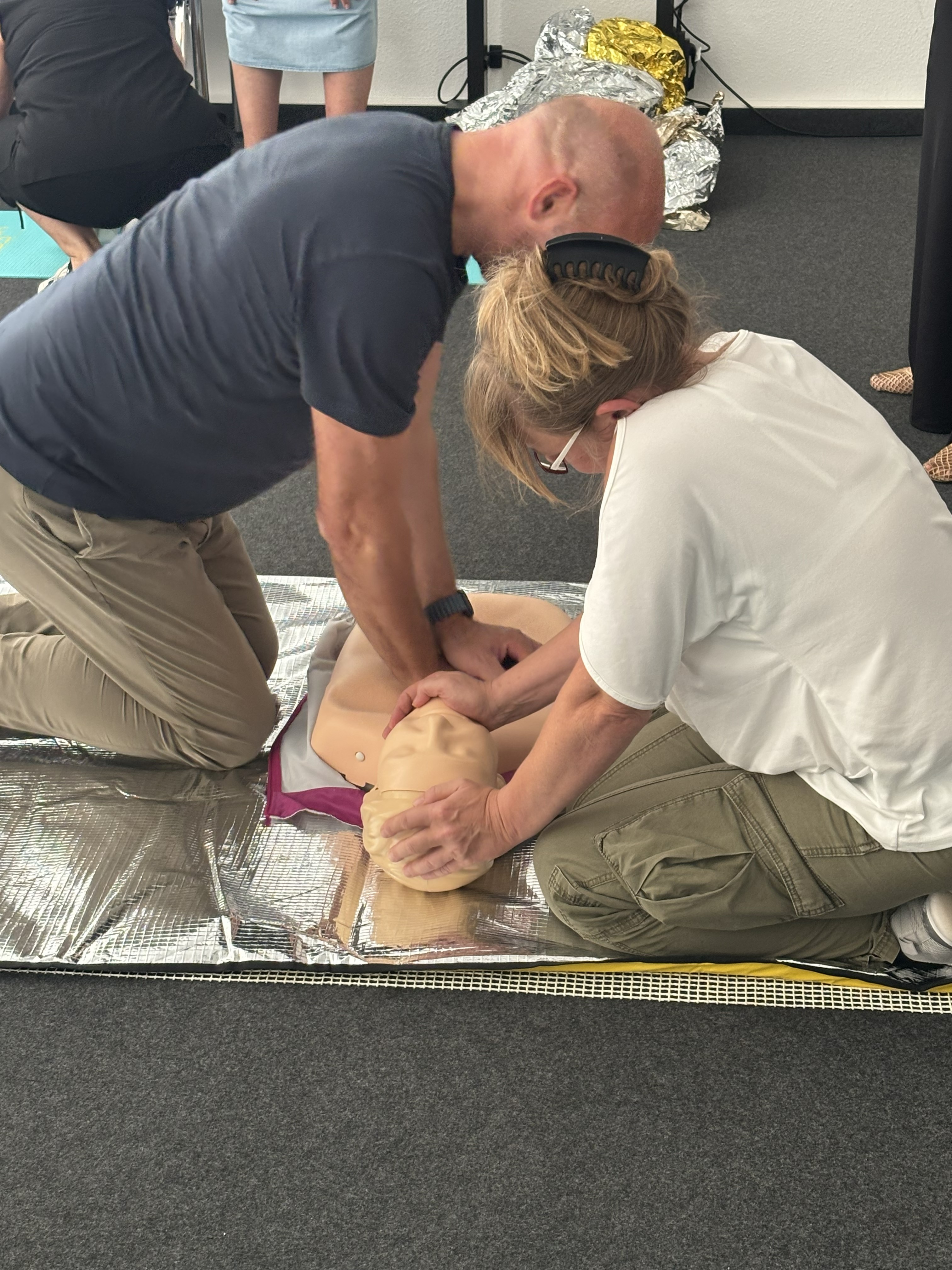 Zwei Teilnehmende üben Erste-Hilfe-Maßnahmen an einer Reanimationspuppe auf einer Rettungsdecke; eine Person führt Herzdruckmassagen durch, während die andere den Kopf stabilisiert.