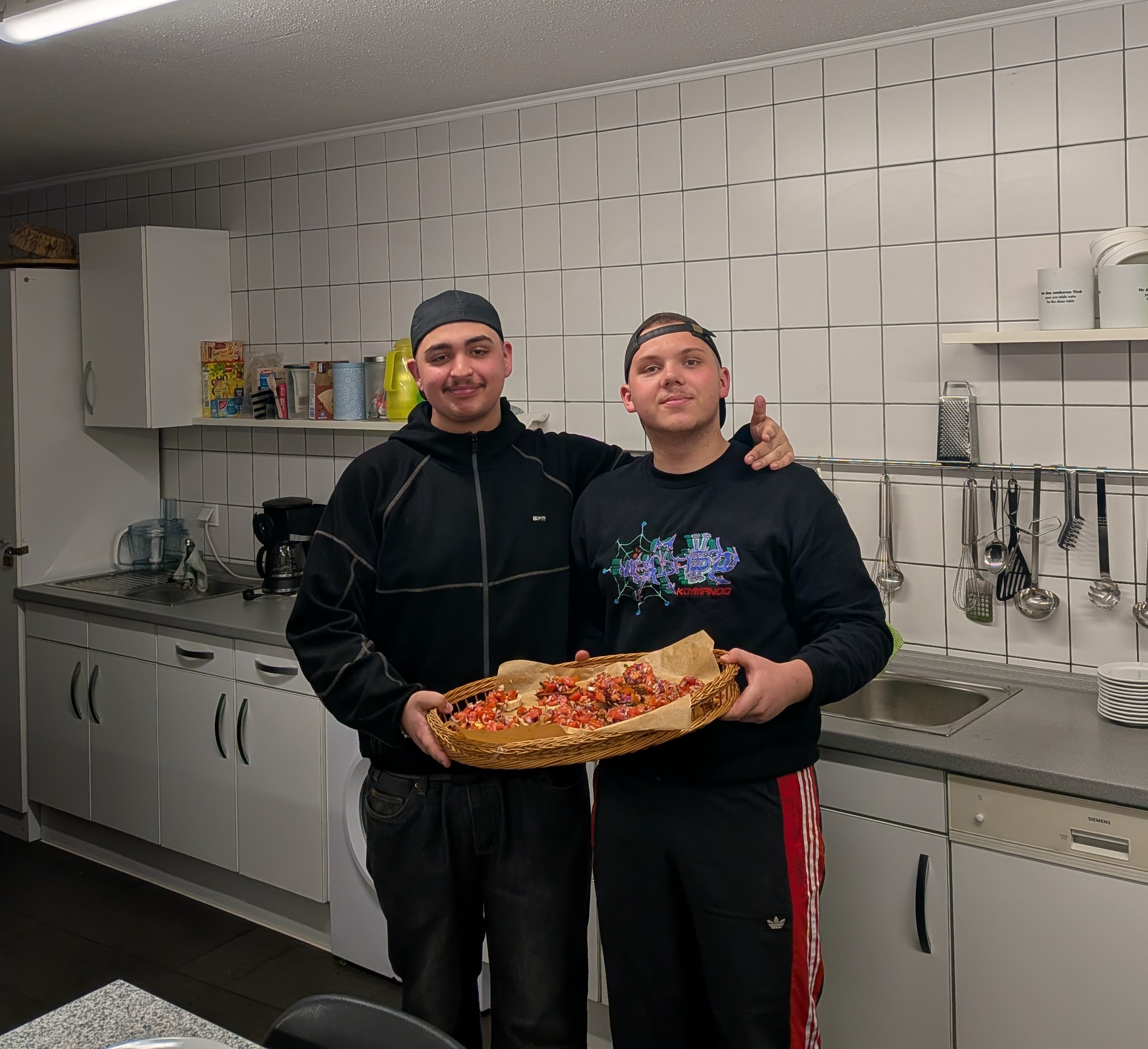 Zwei junge Männer stehen in einer Schulküche und präsentieren gemeinsam ein Tablett mit frisch zubereiteter Bruschetta.
