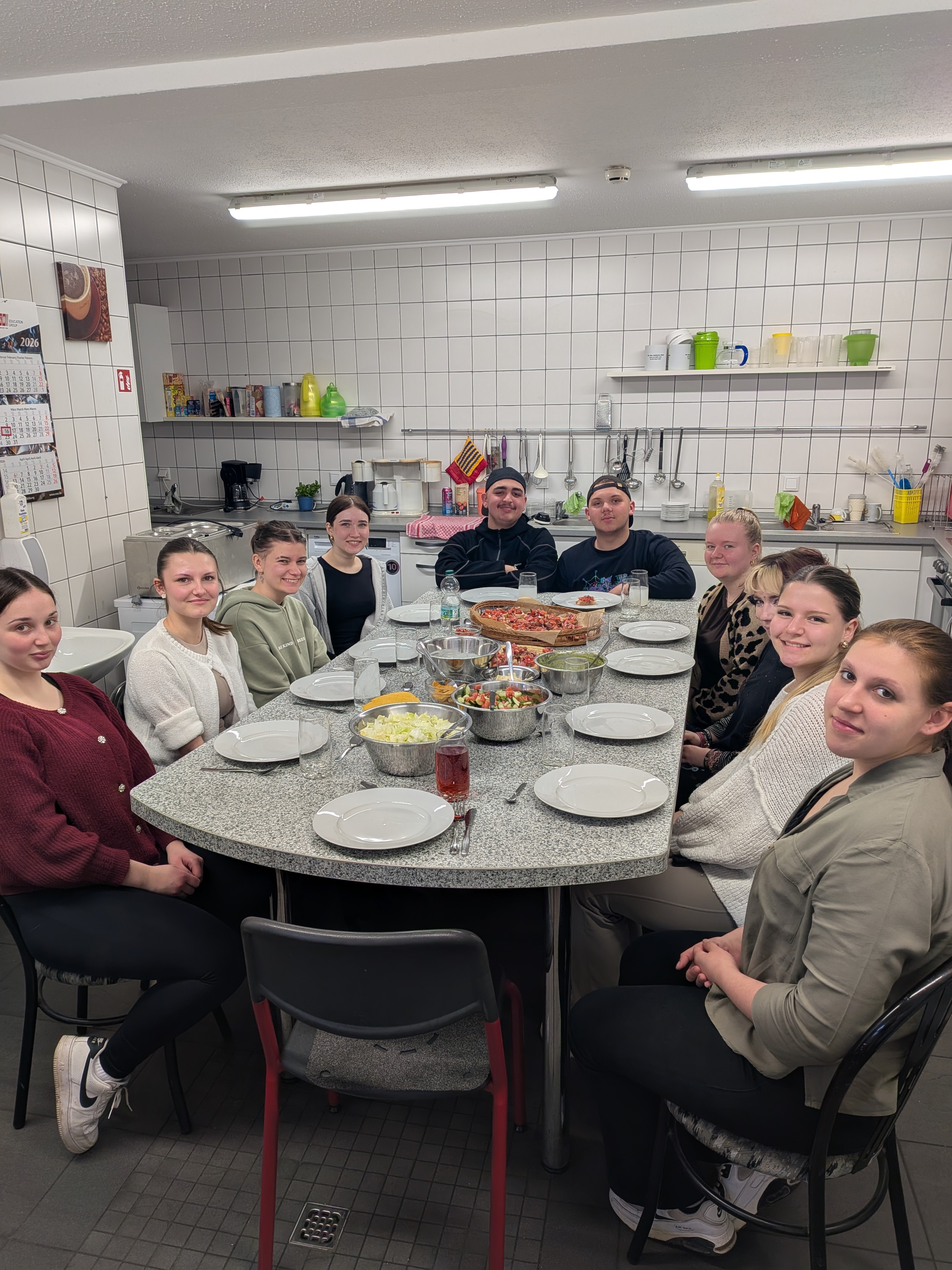 Eine Gruppe von zehn Jugendlichen sitzt in einer Schulküche gemeinsam an einem gedeckten Tisch mit Salaten, Brot und weiteren Speisen.