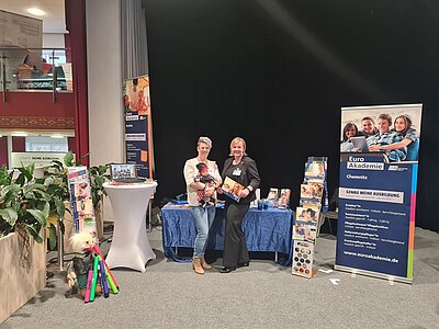 Zwei Mitarbeiterinnen posieren in der Halle am Messestand für ein Foto. Im Hintergrund große Roll-Ups der Euro Akademie, ein Stehtisch, Infobroschüren.