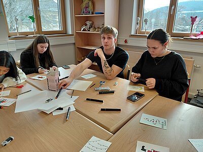 Mehrere Studierende sitzen an einem Tisch und gestalten Weihnachtskarten, ein junger Mann in schwarzem T-Shirt greift nach einem Stift.