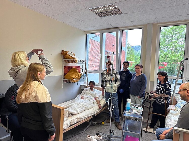 Die Lehrerin mimt eine Patientin, die im Krankenhausbett liegt, umgeben von mehreren Auszubildenden, die neben ihr stehen oder sitzen und für ein Gruppenbild in Richtung eines Smartphones lächeln. Im Raum befinden sich medizinische Geräte, ein Infusionsständer, ein Rollator und ein Servierwagen. Durch die großen Fenster ist ein rotes Gebäude und grüne Bäume zu sehen. Die Atmosphäre wirkt gut gelaunt, ruhig und freundlich.
