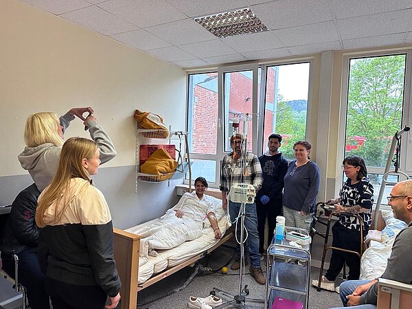 Die Lehrerin mimt eine Patientin, die im Krankenhausbett liegt, umgeben von mehreren Auszubildenden, die neben ihr stehen oder sitzen und für ein Gruppenbild in Richtung eines Smartphones lächeln. Im Raum befinden sich medizinische Geräte, ein Infusionsständer, ein Rollator und ein Servierwagen. Durch die großen Fenster ist ein rotes Gebäude und grüne Bäume zu sehen. Die Atmosphäre wirkt gut gelaunt, ruhig und freundlich.
