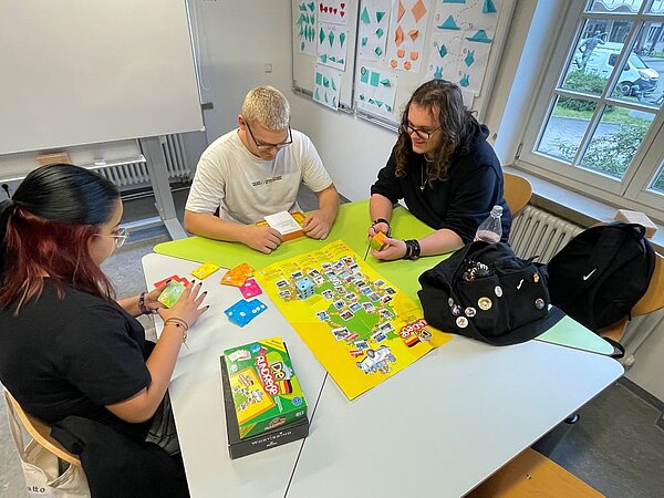 Drei Personen spielen an einem Tisch ein Brettspiel mit gelbem Spielplan; Spielkarten, Würfel und weiteres Spielmaterial liegen bereit, eine Person hält eine grünfarbene Karte. Im Hintergrund ein Whiteboard und Magnettafeln mit bunten Bildern, daneben Rucksäcke und Taschen.