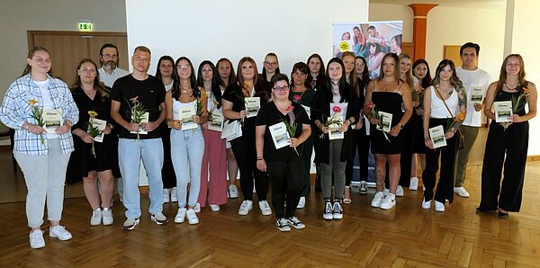 Zu sehen ist eine große Gruppe von Schülerinnen und Schülern, die eine Blume und einen Flyer in der Han halten. Die Gruppe steht in einem Raum mit Parkettboden.