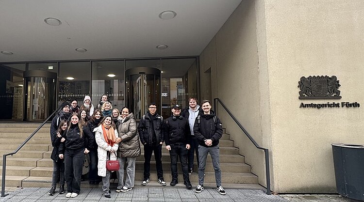 Gruppenfoto von Auszubildenden der Euro Akademie Bamberg vor dem Eingang des Amtsgerichts Fürth. Die Gruppe aus jungen Erwachsenen steht auf den Treppenstufen vor dem Gerichtsgebäude und blickt lächelnd in die Kamera. Viele tragen winterliche Kleidung wie Jacken, Schals und Mäntel. Rechts im Bild ist deutlich das Schild mit der Aufschrift ‚Amtsgericht Fürth‘ an der Außenwand des Gebäudes zu erkennen. Das Foto entstand im Rahmen eines Exkursionsbesuchs, bei dem die Auszubildenden die Ausstellung zu den ‚Nürnberger Prozessen‘ im Amtsgericht Fürth besichtigten und sich mit der historischen Bedeutung von Rechtsprechung, Verantwortung und Demokratie auseinandersetzten.