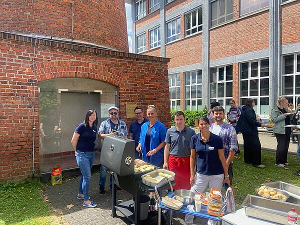 Eine Gruppe steht gemeinsam am Grill im Innenhof vor einem Backsteingebäude, auf dem Grill liegen Würstchen und Grillkäse bereit.