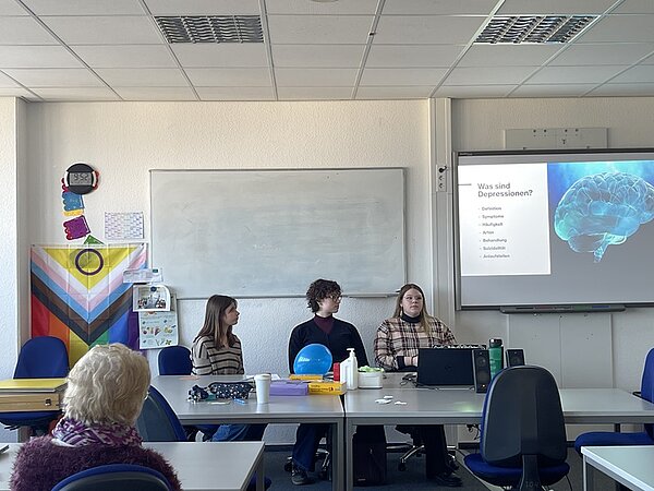 Eine Dreiergruppe von Auszubildenden sitzt in einem Klassenzimmer an einem Tisch und hält eine Präsentation. Die Folie auf dem Bildschirm trägt den Titel „Was sind Depressionen?“ und enthält Stichpunkte zu Definition, Symptomen, Ursachen, Auswirkungen, Behandlung, Suizid und Hilfsangeboten. Auf dem Tisch stehen verschiedene Gegenstände, darunter Luftballons, Becher und ein Laptop. An der Wand hängen eine Regenbogenflagge und einige Blätter Papier. Eine Zuhörerin mit blonden Haaren sitzt im Publikum.