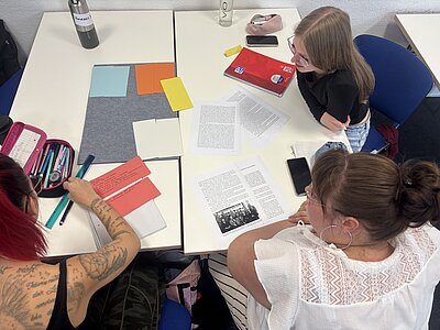 Drei Schülerinnen arbeiten gemeinsam an einem Tisch mit Texten, Notizen und farbigem Bastelmaterial. Sie bereiten Inhalte für ein Präsentationsplakat vor.