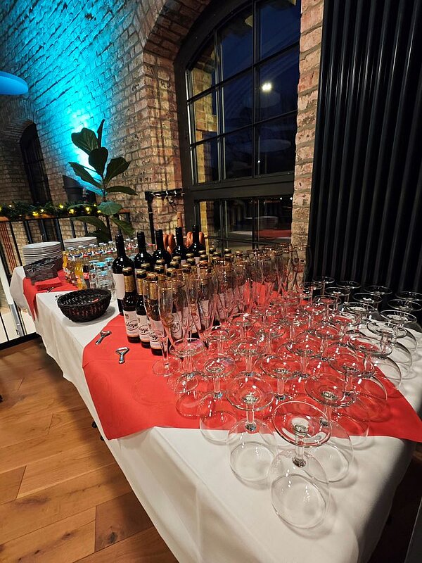 Ein Tisch mit einer Auswahl an Getränken und Gläsern. Auf dem Tisch stehen mehrere Flaschen, darunter Wein- und Sektflaschen, sowie eine Reihe von leeren Weingläsern. Der Tisch ist mit einer weißen Tischdecke und einer roten Tischläufer bedeckt. Im Hintergrund sind Ziegelsteinwände und dekorative Pflanzen zu sehen.