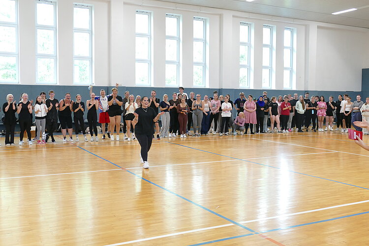 In einer hellen Sporthalle mit hohen Fenstern steht eine Reihe klatschender und jubelnder Personen, die eine junge Frau im Vordergrund anspornen.