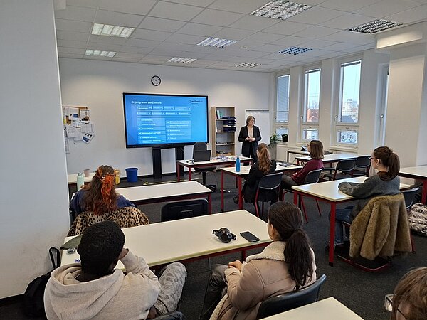 Vortrag an der Euro Akademie Berlin: Eine Referentin des Auswärtigen Amtes steht vorne im Klassenraum und informiert Schülerinnen und Schüler über internationale Karrieremöglichkeiten für Fremdsprachenkorrespondentinnen und Fremdsprachenkorrespondenten. Die Auszubildenden sitzen an Tischen und hören aufmerksam zu. Auf einem großen Bildschirm wird eine Präsentation mit Informationen zu Einstiegsmöglichkeiten, Tätigkeitsfeldern und internationalen Einsatzorten gezeigt. Der helle Unterrichtsraum verfügt über große Fenster, moderne Technik und eine ruhige Lernatmosphäre, die den professionellen Rahmen der Informationsveranstaltung unterstreicht.