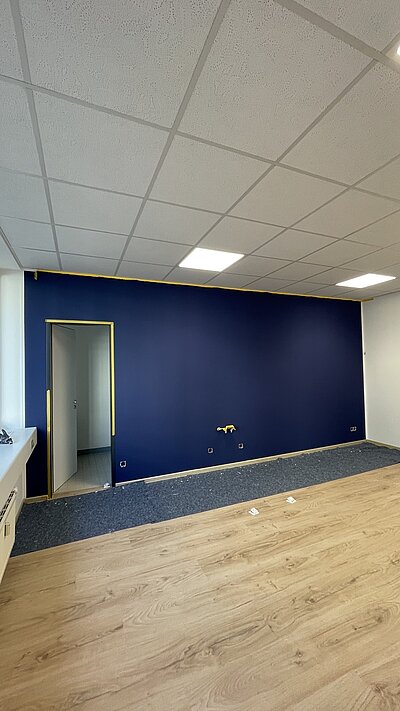 Blick in einen großen, hellen Raum, in dem eine Wand dunkelblau gestrichen ist und Malerfließ ausliegt.