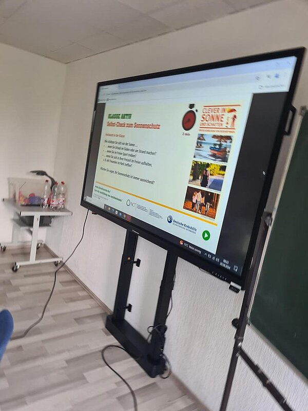 Auszeichnung „Clever in Sonne und Schatten“ 2024 Auf einem großen Bildschirm sieht man Infos zum Selbst-Check zum Sonnenschutz.