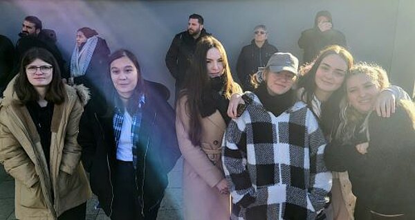 Euro Akademie und Euro-Schulen on Fire! Eine Gruppe von sechs jungen Frauen steht nebeneinander draußen in einer Menschenmenge. Sie tragen warme Kleidung wie Mäntel und Jacken, passend für kühles Wetter. Einige der Frauen haben den Arm um ihre Freundinnen gelegt. Im Hintergrund stehen weitere Personen, die ebenfalls warme Kleidung tragen und teilweise von Sonnenlicht beschienen werden.