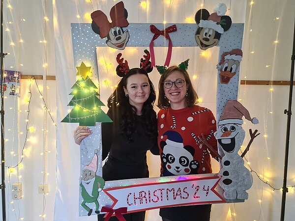Zwei Frauen halten einen weihnachtlichen Fotorahmen hoch, durch den sie in die Kamera blicken und lächeln.