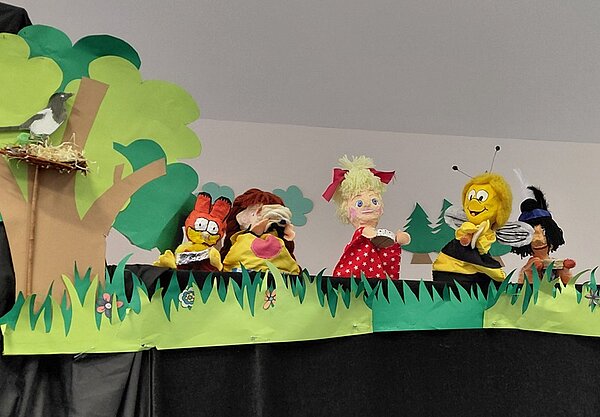 Bühnenbild mit mehreren Handpuppen vor einer Kulisse aus grünem Papier, die Bäume, Gras und Blumen darstellt. Links sitzt ein Vogel in einem Nest auf einem Baum. Die Puppen zeigen verschiedene Figuren, darunter eine Figur mit Brille und roten Haaren, eine Puppe mit blonden Zöpfen und rotem Kleid mit weißen Punkten, eine gelbe Bienenfigur und eine weitere Figur mit dunklen Haaren und Hut. Die Szene wirkt wie ein Puppentheater für Kinder.