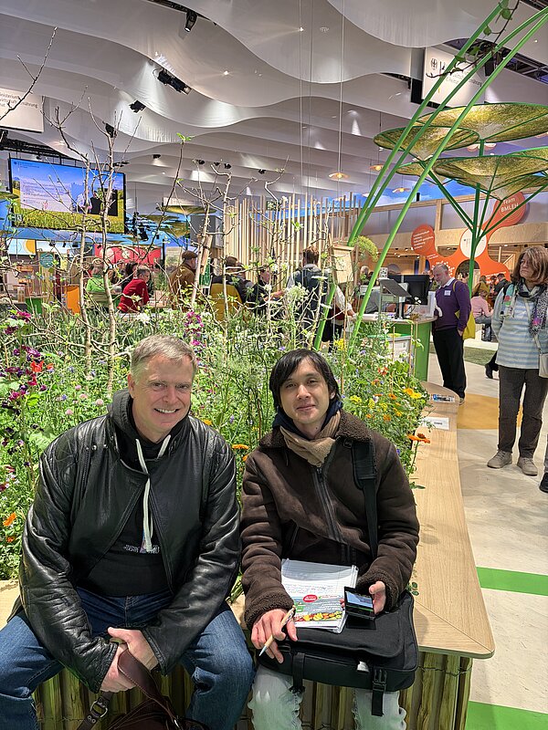 Lehrer und Schüler sitzen vor einem bunten Blumenbeet in einer Messehalle