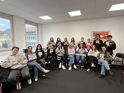 Für ein Gruppenfoto haben sich die Schüler*innen in einer Ecke des Klassenzimmers gesammelt. Sie sitzen und stehen teilweise. Jede*r Schüler*in hält ein Buch bzw. eine Broschüre des Projekts ReSi in die Kamera.