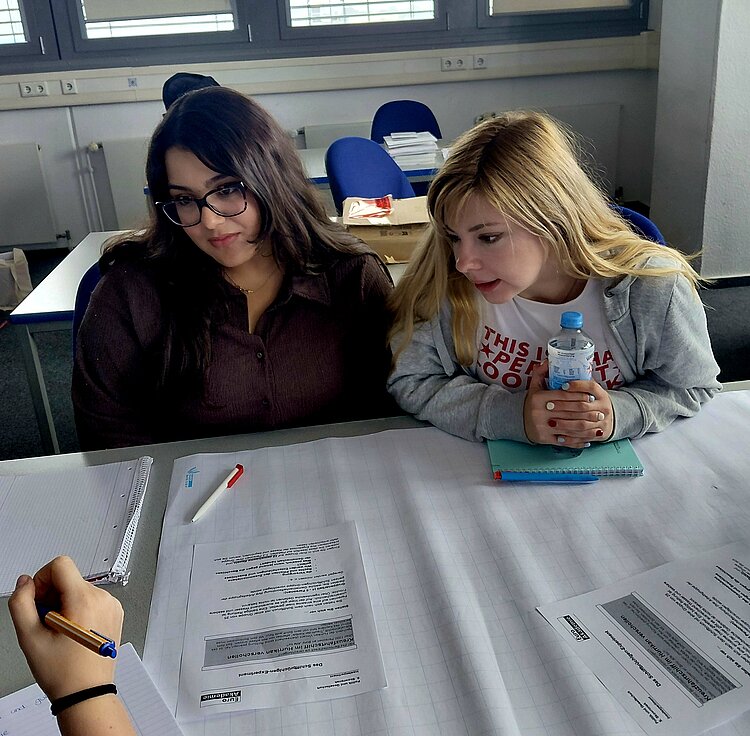 Zwei Schülerinnen sitzen an einem Tisch in einem Klassenzimmer. Sie schauen konzentriert auf Papiere, die vor ihnen liegen. Die rechte Schülerin hält eine Wasserflasche in der Hand. Links im Vordergrund ist die Hand einer weiteren Person zu sehen, die einen Stift hält.