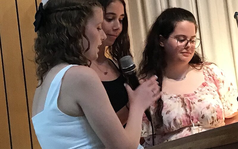 Drei junge Frauen stehen an einem Podium. Eine der Frauen spricht in ein Mikrofon. Alle tragen festliche Kleidung.
