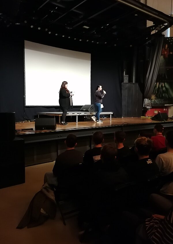 Eine Auszubildende der Euro Akademie Zwickau steht auf der Bühne im Alten Gasometer und moderiert die Schulkino-Vorführung an. Der Raum ist bereits abgedunkelt, im Hintergrund sieht man die große weiße Leinwand. 