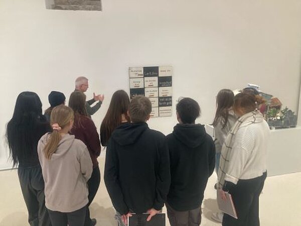 Eine Gruppe von jungen Erwachsenen steht in einer Kunstgalerie und hört einer Person zu, die ihnen ein Kunstwerk an der Wand erklärt.