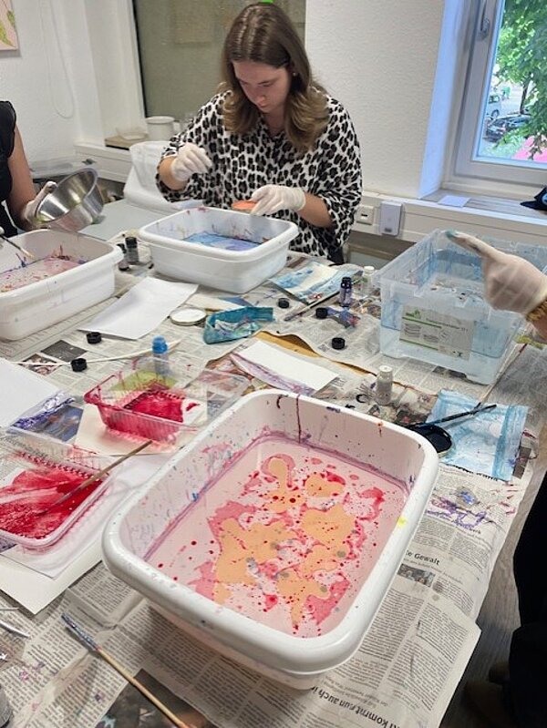 Eine Person in einem gemusterten Oberteil arbeitet an einem Tisch mit mehreren Kunststoffwannen, die mit farbigen Flüssigkeiten gefüllt sind. Sie trägt Handschuhe und konzentriert sich auf ihr Projekt. Im Vordergrund steht eine weiße Kunststoffwanne, die mit roter und rosafarbener und gelber Farbe gefüllt ist.