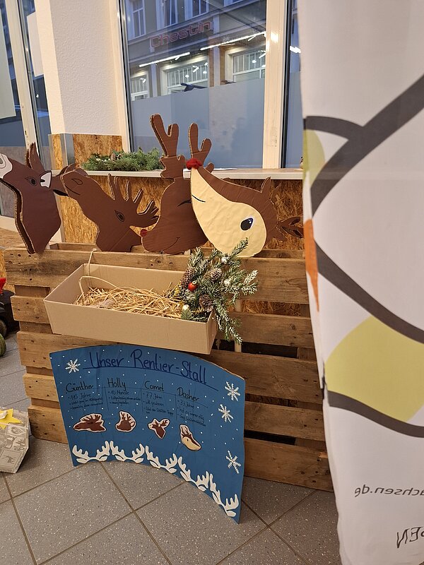 Aus Pappkarton ausgeschnittene Rentiere stecken in einer Holzpalette, davor steht ein blauer Schildkarton mit der Überschrift „Unser Rentier-Stall“ und kurzen Beschreibungen der Tiere.