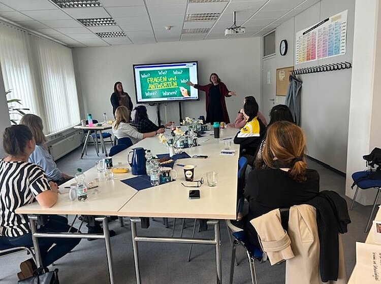 Eine Gruppe von Teilnehmenden sitzt um einen großen Konferenztisch in einem hellen, gut beleuchteten Raum. Zwei Personen stehen vorne neben einem digitalen Display, auf dem eine Folie mit deutschem Text angezeigt wird: „Fragen und Antworten“ sowie Wörter wie „Wer?“, „Wo?“, „Was?“, „Wozu?“, „Warum?“ und „Wann?“. Verschiedene Gegenstände, darunter Wasserflaschen und Becher, stehen herum.