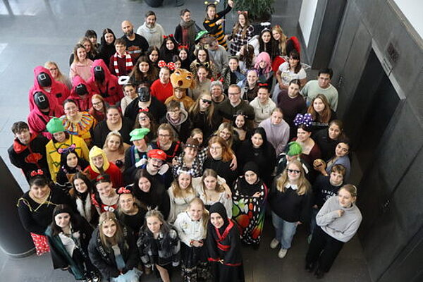 Fasching – Ein Tag voller Spaß und Gemeinschaft Eine große Gruppe verkleideter Schüler*innen mit ganz unterschiedlichen Kostümen steht im Eingangsbereich eines Gebäudes, vor einem Fahrstuhl. Die Personen blicken nach oben, wo offenbar die Person steht, die das Foto aufnimmt.