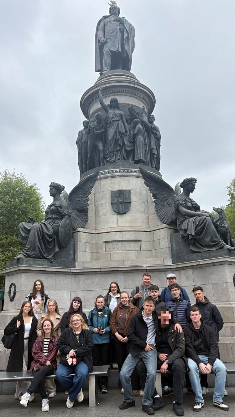 Gruppe von jungen Erwachsenen vor einem großen Denkmal mit Statuen; einige sitzen auf einer Bank, andere stehen dahinter.