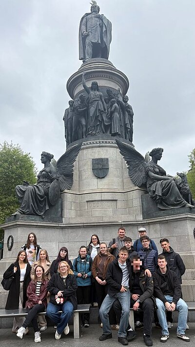 Gruppe von jungen Erwachsenen vor einem großen Denkmal mit Statuen; einige sitzen auf einer Bank, andere stehen dahinter.