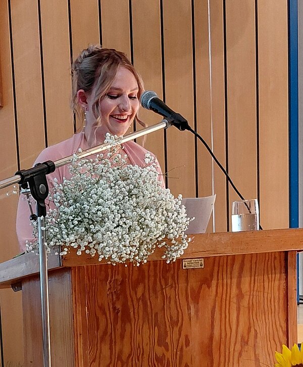 Absolventin steht am Rednerpult und spricht lächelnd ins Mikrophon. Auf dem Pult steht ein Glas Wasser und liegen Blumen.