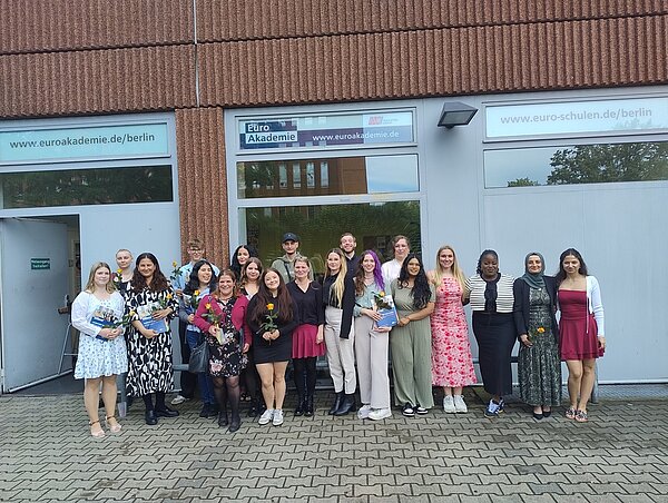 Vor dem Gebäude der Euro Akademie Berlin haben sich die Absolvent*innen für ein Foto positioniert. Sie tragen festliche Kleidung und halten teilweise eine gelbe Rose sowie blauen Mappen der Euro Akademie in den Händen.
