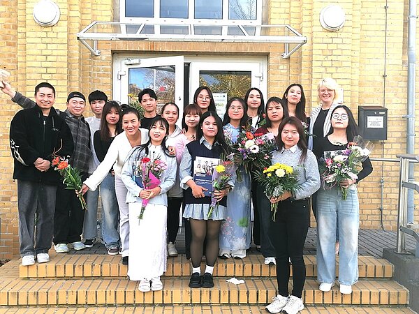 Die Absolventinnen und Absolventen der Pflegefachkraft-Ausbildung an der Euro Akademie Bitterfeld-Wolfen stehen vor dem Eingangsbereich des Schulgebäudes. Die Gruppe hält bunte Blumensträuße und posiert gut gelaunt für ein Foto zur feierlichen Zeugnisübergabe.