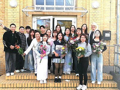 Die Absolventinnen und Absolventen der Pflegefachkraft-Ausbildung an der Euro Akademie Bitterfeld-Wolfen stehen vor dem Eingangsbereich des Schulgebäudes. Die Gruppe hält bunte Blumensträuße und posiert gut gelaunt für ein Foto zur feierlichen Zeugnisübergabe.