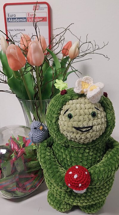 Im Vordergrund steht die grüne, gehäkelte Stofffigur Moosi mit freundlichem Gesichtsausdruck. Die Figur hält einen roten Fliegenpilz in der Hand und trägt eine gehäkelte Blume auf dem Kopf. Auf der linken Schulter sitzt ein kleiner, gehäkelter Vogel. Im Hintergrund stehen zwei Glasvasen: Eine enthält künstliche Tulpen, die andere ist mit bunten Papierstreifen gefüllt. Im oberen Bereich ist ein Schild mit der Aufschrift 'Euro Akademie Schulen' und einem weiteren Namen zu sehen.