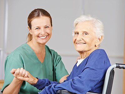 Eine Pflegekraft in grüner Berufsbekleidung und eine ältere Frau in einem blauen Oberteil sitzen freundschaftlich beieinander. Die ältere Frau sitzt in einem Rollstuhl, während die Pflegekraft ihre Hand hält. Beide lächeln herzlich und wirken glücklich. Der Hintergrund zeigt einen neutralen, hellen Raum, der eine warme und vertrauensvolle Atmosphäre vermittelt.